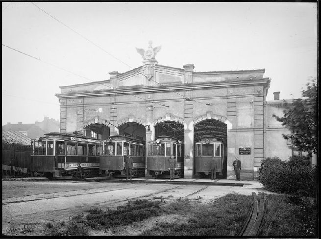 Unikátní snímek původní vozovny elektrické dráhy v prostoru dnešních ústředních dílen Dopravních podniků v Cukrovarské ulici. Ve vratech remízy vidíme (zleva) motorové vozy evid. č. 18, 8, 12 a 23. Původní čtyřkolejná remíza byla zděná budova se spouštěcími roletovými vjezdovými vraty. Dřevěná střecha byla nesena nýtovanou konstrukcí z válcovaných profilů. Koleje remízy byly přístupné pouze přes krátkou přesuvnu (na ruční pohon). V hale vozovny nebyla trolej – vozy musely být přesouvány zřejmě ručně. Tato původní malá remíza byla později zbourána a na jejím místě vyrostly dnešní ústřední dílny. Nahrazena byla novou vozovnou, která stojí v Cukrovarské ulici dodnes a je využívána pro trolejbusy. Na snímku je velmi zajímavá orientační tabulka ukazující směr ke kanceláři, která opět dokládá „českost“ Plzně v období Rakouska – Uherska. Na sloupu vrat mezi vozy evid. č. 12 a 23 je černá tabulka s křídou napsanými čísly – pravděpodobně jde o informační tabulku o umístění vozů ve vozovně.
Skleněný negativ formátu 18 × 24 cm  je uložen ve sbírkách ZČM, sign. NMP 78. Autor neznámý.