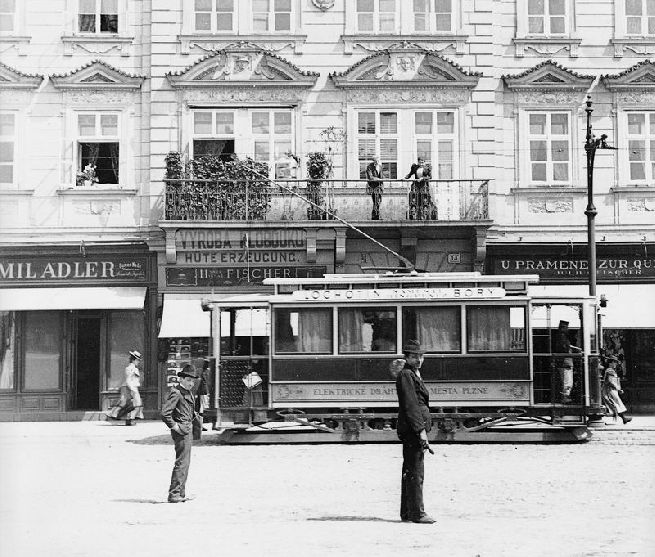 Motorový vůz druhé výrobní série evid. č. 21—25, podle větracího nástřešku však můžeme evid. č. 21 vyloučit. Snímek dobře ukazuje základní rozdíly druhé série – síťovaná nástupní dvířka, schodnice se zády z děrovaného plechu, jiné zavěšení ochranného rámu, který nepřesahuje půdorysný profil vozu, čelní traverza, hlavní pojistka na opačné straně vozu, jiné zavěšení tlapových motorů na podvozku. Vůz stojí na náměstí u domu č. p. 14, který má dnes pozměněnu partii v místě balkónu, který byl později obestavěn a zasklen. Na balkóně vidíme patrně místní honoraci – dáma přísně kontroluje situaci na ulici. Na obchodech vidéme české nápisy, což je důkaz pro konstatování, že Plzeň byla skutečně česká. Snímek byl pořízen v prvních letech provozu, pravděpodobně v roce 1900. Autor neznámý. Sbírky ZČM, sign. 2287. Skleněný negativ 13×18 cm. Výřez.